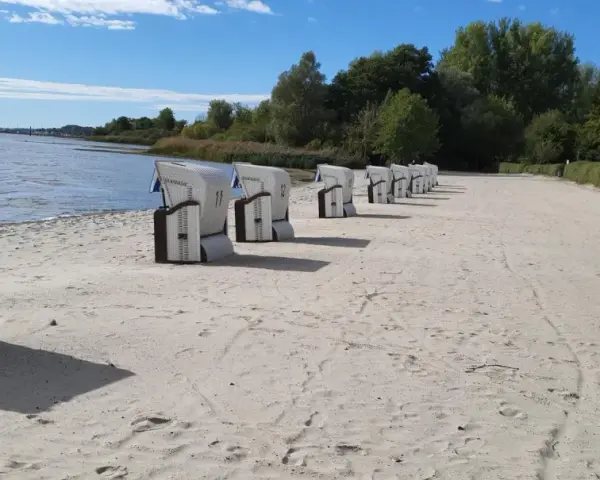 Reihe von Strandkörben am klink strand. Das Wasser glitzert im Hintergrund, Bäume säumen den Horizont.