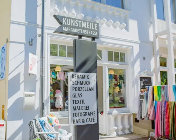 Schild an der Kunstmeile Binz: Keramik, Schmuck, Porzellan, Glas, Textil, Malerei, Fotografie, Bar & Café. Bunte Schals hängen davor.