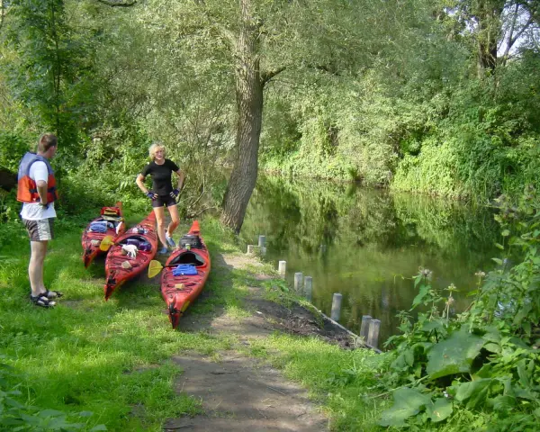 Am Ufer der Kanustation Mirow stehen drei rote Kajaks bereit. Ein Mann mit Schwimmweste und eine Frau in Sportkleidung warten auf ihre Paddeltour.