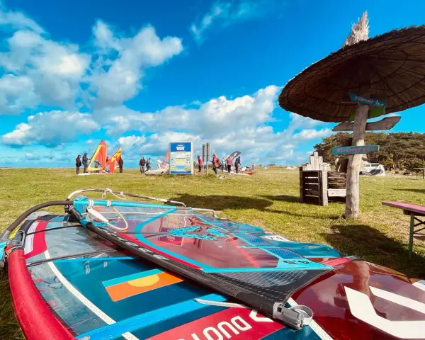 Windsurfausrüstung liegt am ummanz strand. Im Hintergrund bunte Segel und Menschen, die sich auf das Wasser vorbereiten.