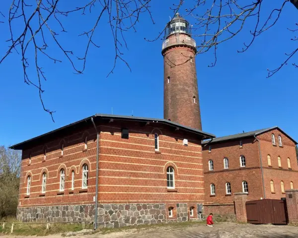 Der Leuchtturm Prerow ragt imposant in den blauen Himmel. Ziegelbauten mit Rundbogenfenstern säumen seinen Fuß.