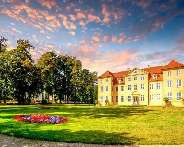 Gelbes Schloss mit roten Dächern, umgeben von grünen Bäumen und Blumenbeeten. Ein malerischer Anblick auf der Liebesinsel Mirow.