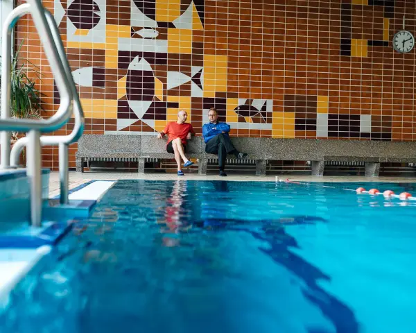 Zwei Männer sitzen in der Schwimmhalle Anklam auf einer Bank vor einer bunten Kachelwand mit Fischmotiven.