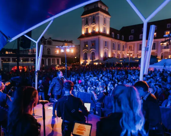 Konzert mit Sänger und Band auf der Bühne, beleuchtet von blauen Lichtern. Im Hintergrund die beleuchtete Architektur und eine Menschenmenge bei Veranstaltungen am Binz Kurplatz.