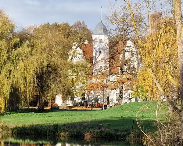 Herbstliche Farben umgeben das Wasserschloss Quilow. Ein Teich spiegelt die Bäume und das weiße Gebäude mit seinem markanten Turm.