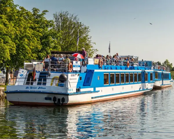 Fahrgastschifffahrt Wichmann: Zwei blau-weiße Schiffe mit Passagieren auf dem Wasser, umgeben von grünen Bäumen und klarem Himmel.