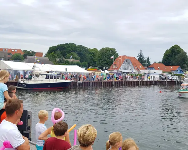 Menschen genießen das bunte Treiben beim Rock am Hafen. Boote liegen im Wasser, ein Rettungsboot fährt vorbei.
