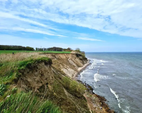 Steilküste mit grasbewachsenem Abhang, der zum steinbeck Strand und dem Meer abfällt. Wolken ziehen über den blauen Himmel.