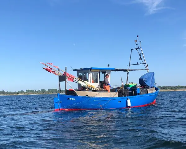 Blaues Fischerboot "RERIK" mit roten Markierungen auf dem Wasser. Ein Mann steht an Deck, rote Fahnen ragen empor.