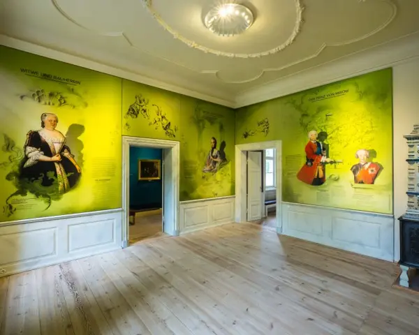 Ein Raum im Schloss Mirow mit Wandmalereien, die historische Persönlichkeiten und Karten zeigen. Ein Kachelofen steht rechts.