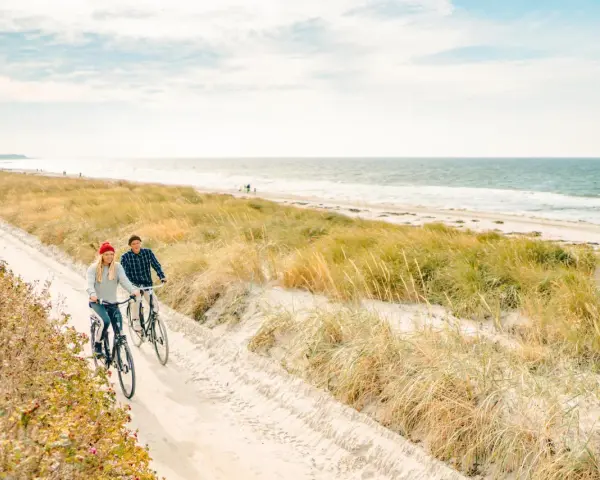 Ein Paar genießt seine baltischen Abenteuer auf Fahrrädern entlang eines sandigen Weges am Meer, umgeben von Dünen und Gräsern.