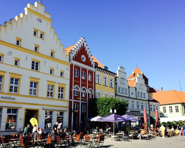 Bunte Häuserfronten säumen den mv markt. Menschen sitzen in Cafés unter Sonnenschirmen und genießen den sonnigen Tag.