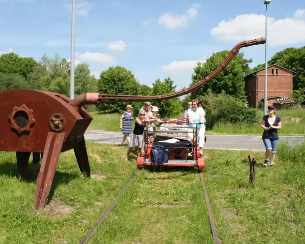 Eine Gruppe von Menschen auf einer Draisine Dargun, neben einer rostigen Skulptur.
