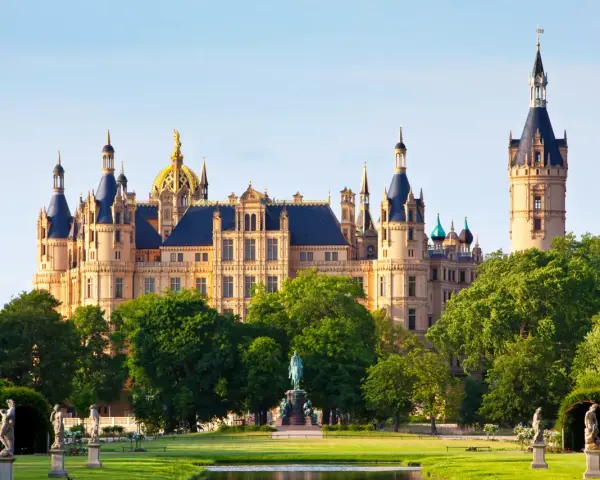 Das Schweriner Schloss, ein Märchenschloss mit goldenen Kuppeln und Türmen, thront majestätisch über einem grünen Park mit Statuen und einem Teich.