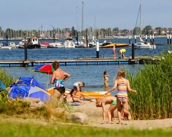 Familien genießen einen sonnigen Tag am Wasser. Kinder planschen, während Boote im Hintergrund ankern. Ein perfekter Ort für Seekamp Camping.