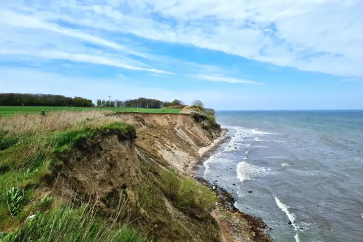 Steilküste mit grasbewachsenem Abhang, der zum steinbeck Strand und dem Meer abfällt. Wolken ziehen über den blauen Himmel.