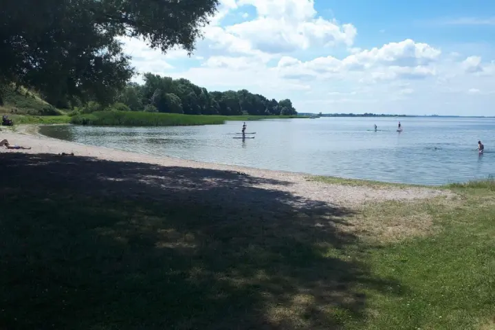 Sonniger Tag am Kummerower See. Menschen entspannen am Strand, andere paddeln auf SUPs. Herrlicher See- und Waldblick.