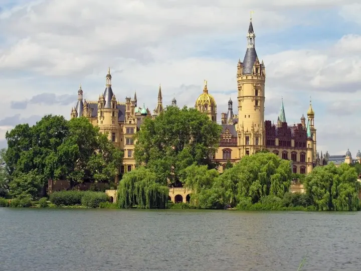 Ein märchenhaftes Schloss mit vielen Türmen und goldenen Kuppeln, umgeben von grünen Bäumen, spiegelt sich im Wasser. Ein gut gremmelin-artiger Anblick.
