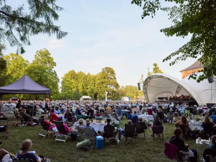 Großes Publikum bei einem Open-Air-Konzert im Rahmen eines **Festivals Mecklenburg-Vorpommern**. Eine Bühne mit Orchester, viele Sitzende auf Rasenflächen.