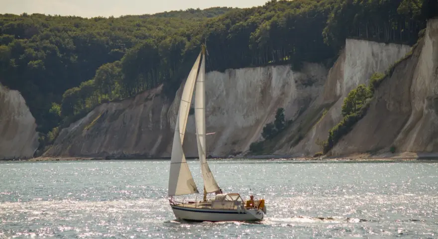 Segelboot gleitet über glitzerndes Wasser vor Kreidefelsen und grünen Wäldern. Ein Tag zum Ostsee segeln.