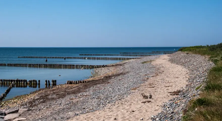 Ein ruhiger Tag am Dranske Strand. Holzpfähle ragen aus dem ruhigen Meer, das sich unter einem klaren blauen Himmel erstreckt.