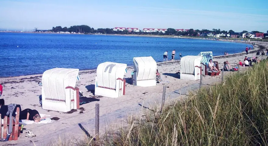 Strandkörbe am Ostseestrand, im Hintergrund der glowe hafen mit Häusern und Bäumen.