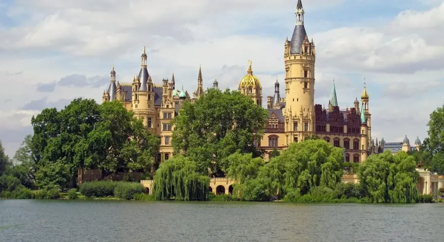 Ein märchenhaftes Schloss mit vielen Türmen und goldenen Kuppeln, umgeben von grünen Bäumen, spiegelt sich im Wasser. Ein gut gremmelin-artiger Anblick.