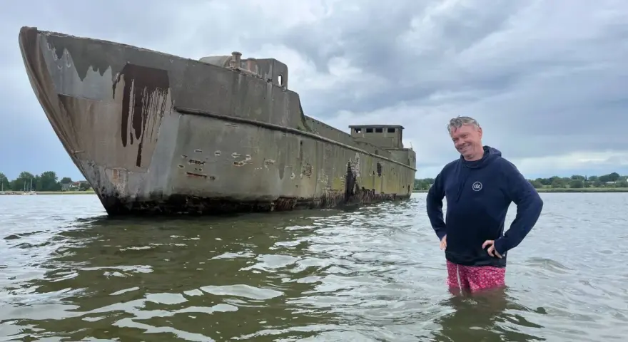 Mann steht im Wasser neben einem verwitterten Betonschiff in Wismar.