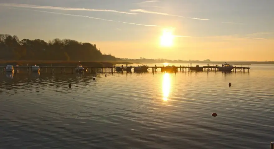 Sonnenaufgang am **Stralsund Strand**. Boote liegen am Steg, die Sonne spiegelt sich im ruhigen Wasser.