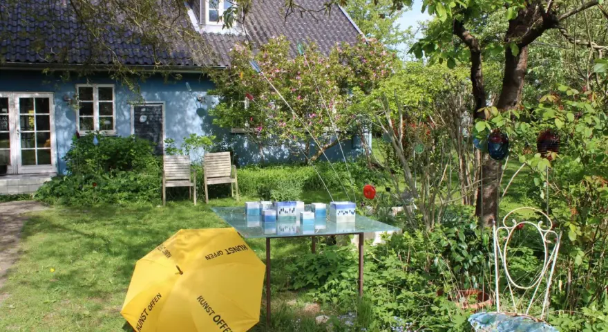 Ein Garten mit blauem Haus, gelbem Regenschirm (Kunst Offen) und Glaskunst auf einem Tisch. Pfingsten 2026 MV.
