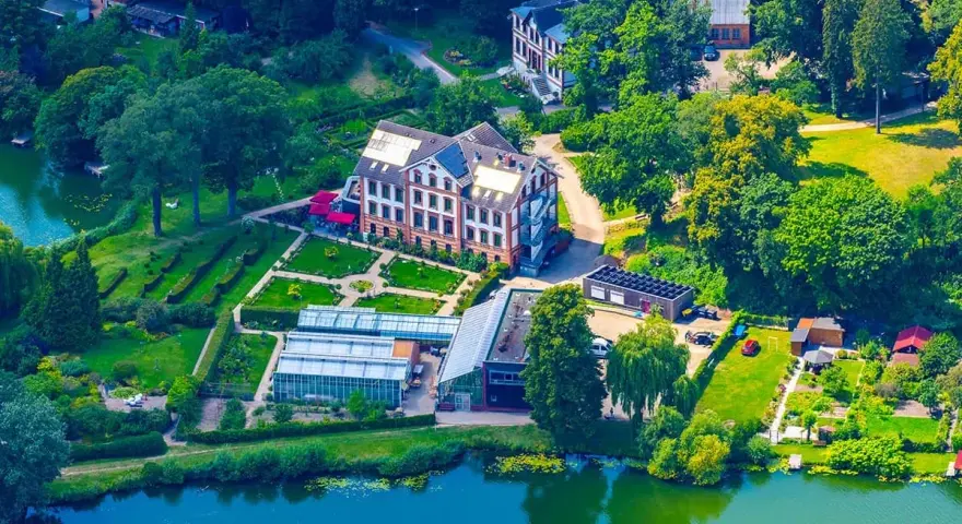 Luftaufnahme des schaugarten am tiefwarensee mit Gewächshäusern, einem Hauptgebäude und gepflegten Gärten am Ufer.