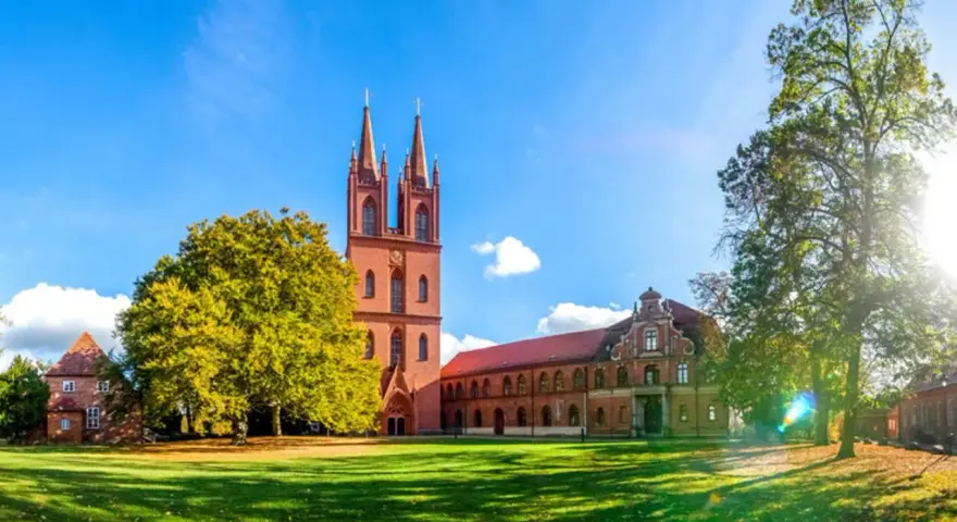Das Kloster Dobbertin erstrahlt im Sonnenschein. Ein imposanter Kirchturm mit zwei Spitzen ragt empor, umgeben von grünen Rasenflächen und alten Bäumen.