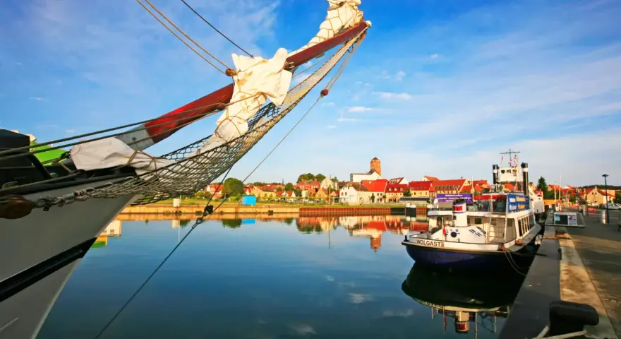 Ein Segelschiff im Vordergrund, im Hintergrund die Stadt Wolgast mit roten Dächern und einem Turm. Ein kleines Boot mit dem Namen "Wolgast" liegt am Kai.