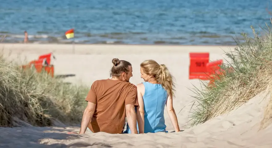 Ein Paar sitzt im Sand am Trassenheide Strand, blickt sich an. Im Hintergrund das Meer und rote Strandkörbe.
