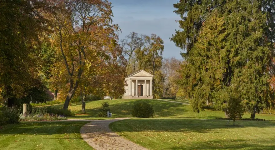 Herbstlicher Spaziergang im Schlossgarten Neustrelitz. Ein Weg führt zu einem antiken Mausoleum, umgeben von bunten Bäumen.