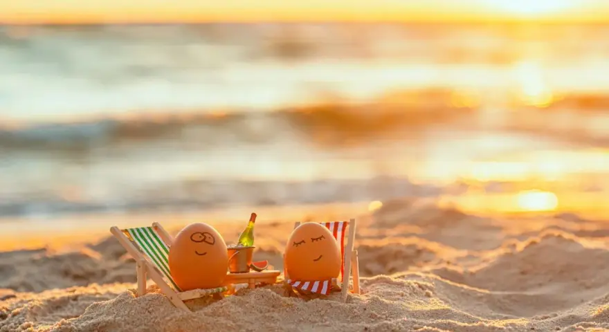 Zwei bemalte Eier entspannen sich in Liegestühlen am Strand bei Sonnenuntergang. Eine Flasche Sekt steht bereit. Eine süße Idee für Veranstaltungen an Ostern.