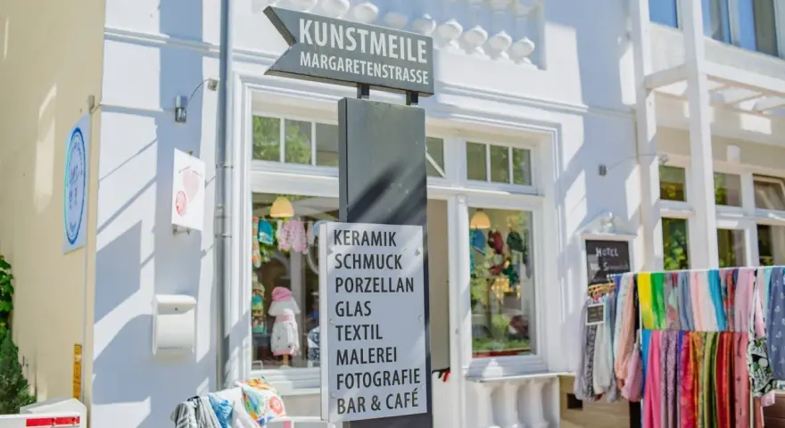 Schild an der Kunstmeile Binz: Keramik, Schmuck, Porzellan, Glas, Textil, Malerei, Fotografie, Bar & Café. Bunte Schals hängen davor.