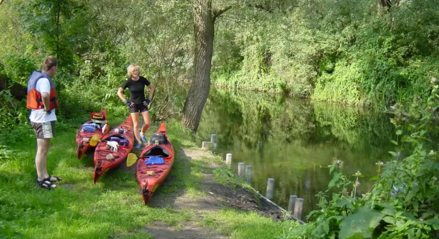 Am Ufer der Kanustation Mirow stehen drei rote Kajaks bereit. Ein Mann mit Schwimmweste und eine Frau in Sportkleidung warten auf ihre Paddeltour.