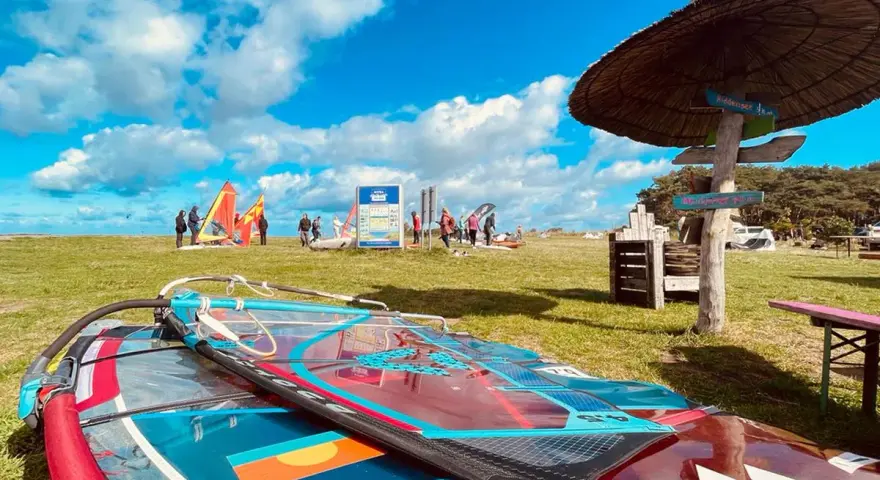 Windsurfausrüstung liegt am ummanz strand. Im Hintergrund bunte Segel und Menschen, die sich auf das Wasser vorbereiten.