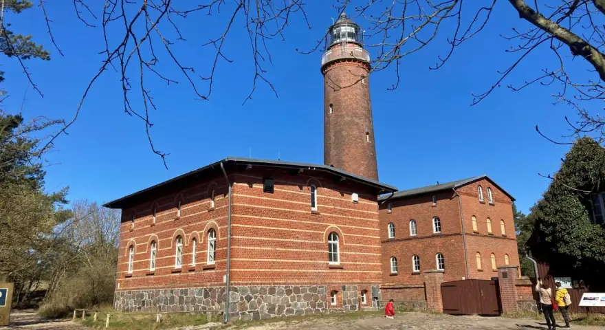 Der Leuchtturm Prerow ragt imposant in den blauen Himmel. Ziegelbauten mit Rundbogenfenstern säumen seinen Fuß.
