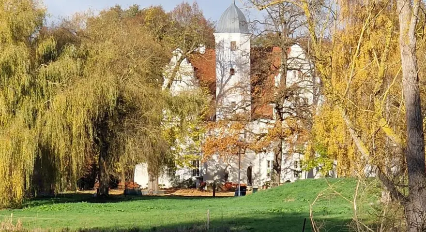 Herbstliche Farben umgeben das Wasserschloss Quilow. Ein Teich spiegelt die Bäume und das weiße Gebäude mit seinem markanten Turm.
