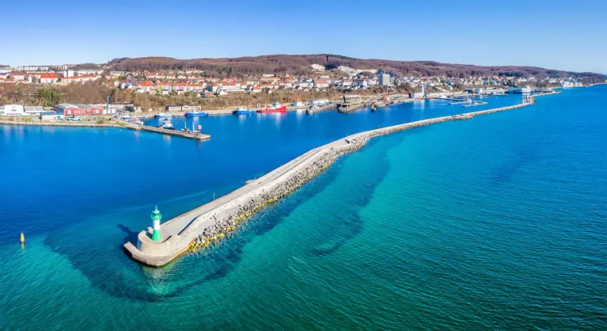 Leuchtturm am Ende einer Mole, die in das klare Wasser vor Sassnitz führt. Boote und die Stadt im Hintergrund.