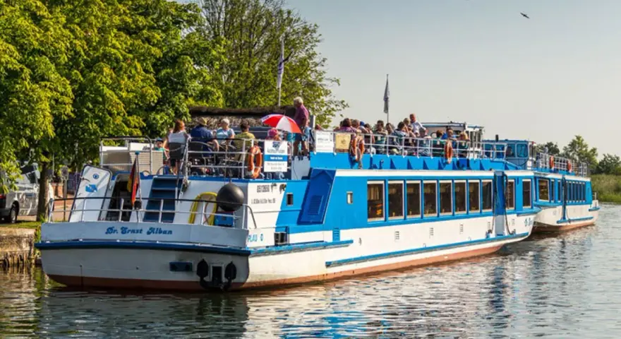 Fahrgastschifffahrt Wichmann: Zwei blau-weiße Schiffe mit Passagieren auf dem Wasser, umgeben von grünen Bäumen und klarem Himmel.