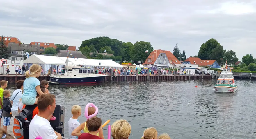 Menschen genießen das bunte Treiben beim Rock am Hafen. Boote liegen im Wasser, ein Rettungsboot fährt vorbei.