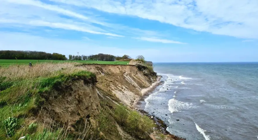 Steilküste mit grasbewachsenem Abhang, der zum steinbeck Strand und dem Meer abfällt. Wolken ziehen über den blauen Himmel.