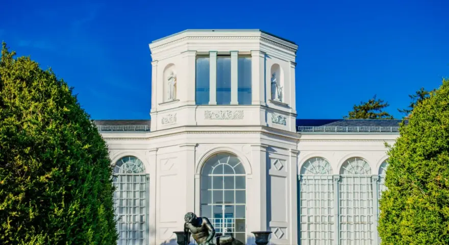 Die Orangerie Putbus, ein strahlend weißes Gebäude mit Skulpturen und großen Fenstern, wird von üppigem Grün eingerahmt.
