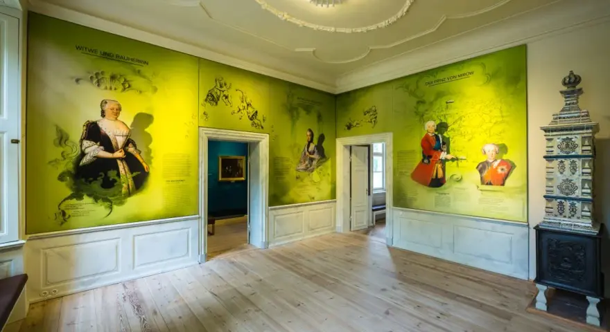 Ein Raum im Schloss Mirow mit Wandmalereien, die historische Persönlichkeiten und Karten zeigen. Ein Kachelofen steht rechts.