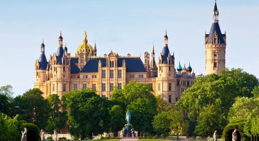 Das Schweriner Schloss, ein Märchenschloss mit goldenen Kuppeln und Türmen, thront majestätisch über einem grünen Park mit Statuen und einem Teich.