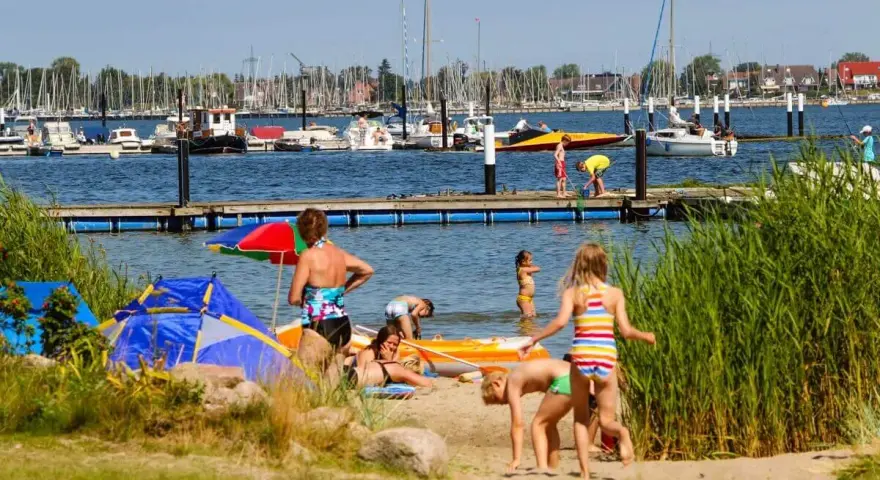 Familien genießen einen sonnigen Tag am Wasser. Kinder planschen, während Boote im Hintergrund ankern. Ein perfekter Ort für Seekamp Camping.