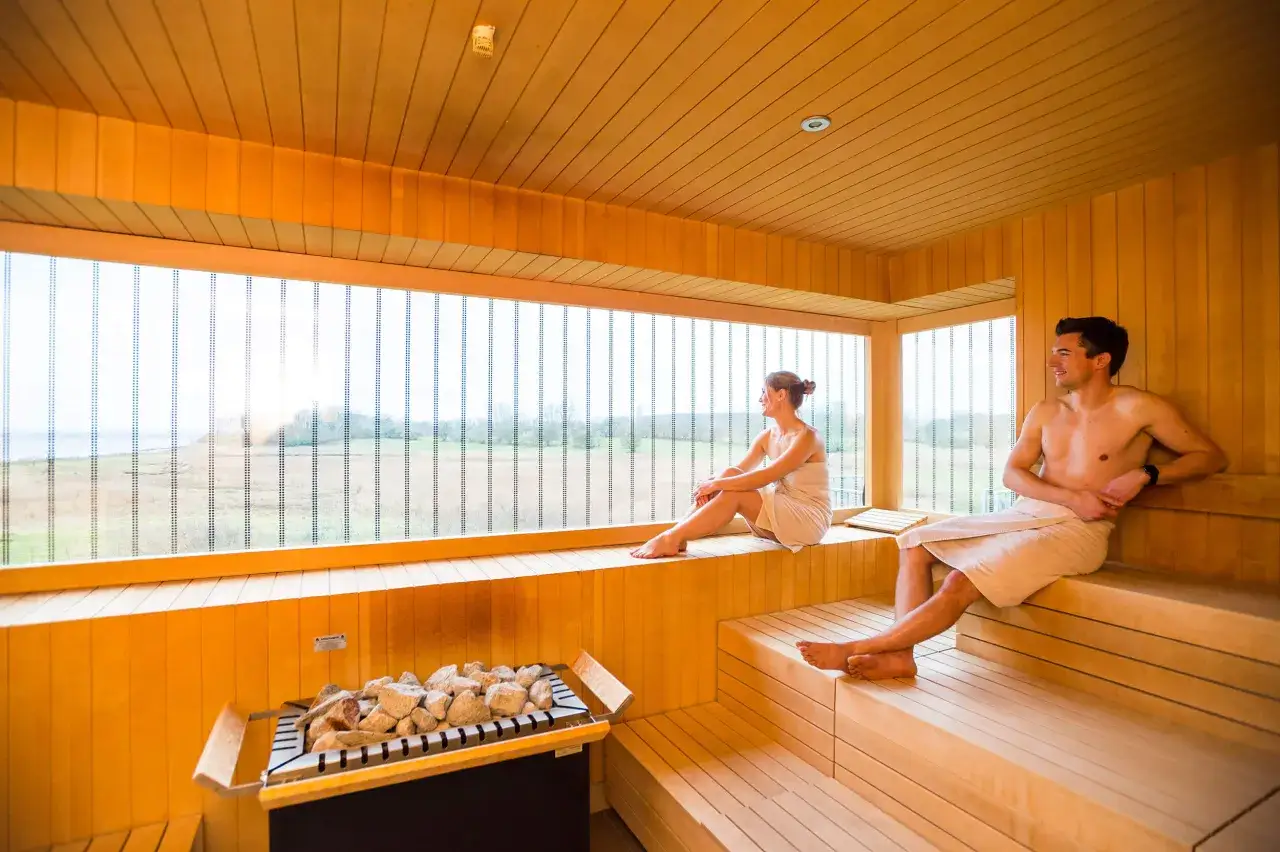 Ein Paar entspannt in einer modernen Therme an der Ostsee. Die holzverkleidete Sauna mit gro&szlig;em Fenster bietet Blick auf die Natur.